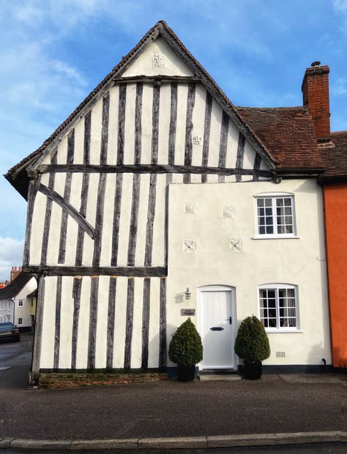 Front view of a traditional English cottage with a distinctive black and white timber-framed facade and white plaster infill. The house features a pitched roof with dark tiles, a white front door flan