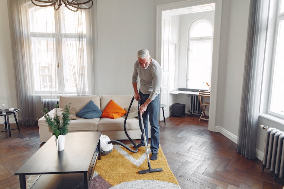 A person is kneeling on a decorative patterned carpet in a residential setting, operating a yellow and black vacuum cleaner for surface cleaning. The individual is wearing blue jeans and a green top, 